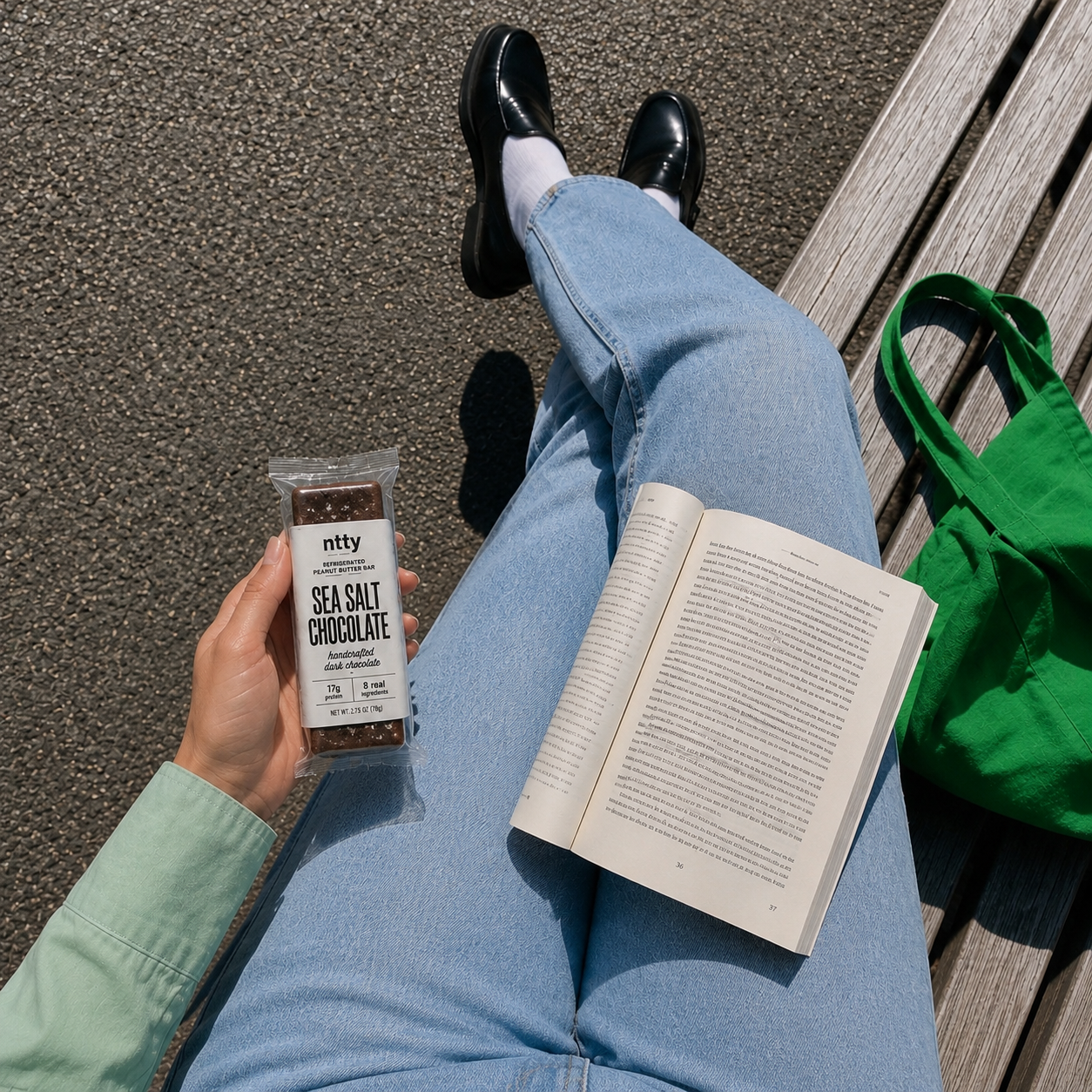 Person reading a book on a bench while holding a ntty Sea Salt Chocolate refrigerated peanut butter bar.
