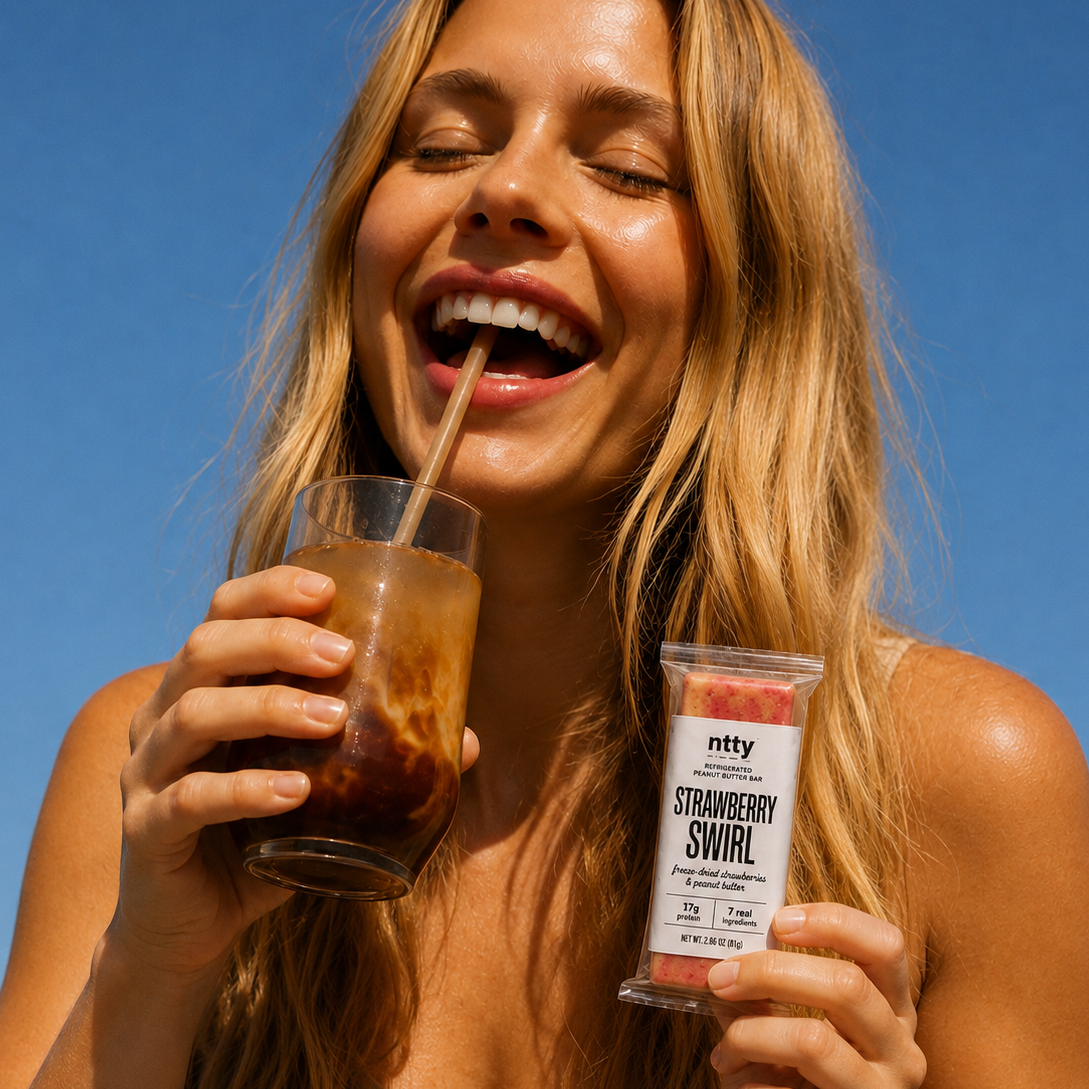 Woman smiling while holding a ntty Strawberry Swirl refrigerated peanut butter bar and an iced coffee outdoors.